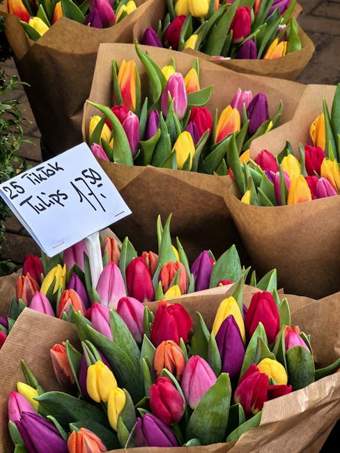 Multiple bouquets of fresh tulips with vibrant pink, yellow, purple, and orange blooms, wrapped in brown kraft paper, are displayed in a floral market setting. The tulips have sturdy green stems and s