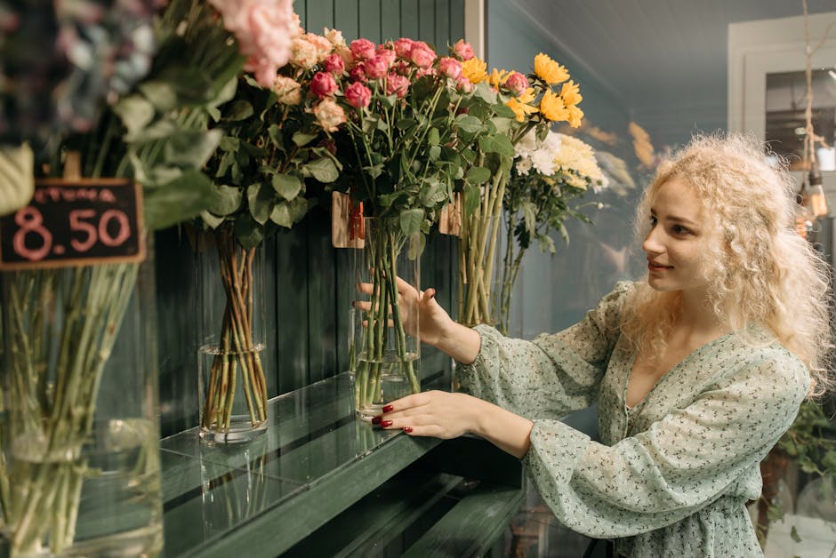 A young woman with curly blonde hair and fair skin selecting a bouquet of fresh pink, yellow, and white flowers from a display of various floral arrangements in a florist shop. She is gently touching 