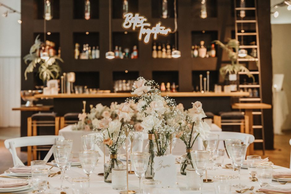 A table setting featuring a floral centerpiece of soft pink and white roses, baby's breath, and greenery arranged in small glass vases. The flowers appear fresh with delicate petals and green stems, p