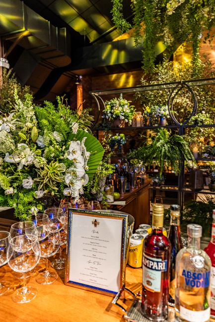 A vibrant floral arrangement featuring white orchids, green foliage, and variegated leaves in a lush, abundant style sits on a wooden bar counter, surrounded by empty wine glasses and bottles of spiri