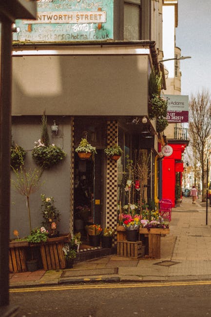 A floral arrangement display in front of a botanical shop on a street corner, featuring a variety of fresh flowers in vibrant pinks, purples, and greens, arranged in wooden crates and pots. The flower