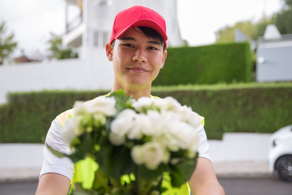 A delivery person in a red cap and high-visibility vest holding a bouquet of fresh white roses and green foliage, extending the floral gift towards the camera outdoors on a residential street. The bou