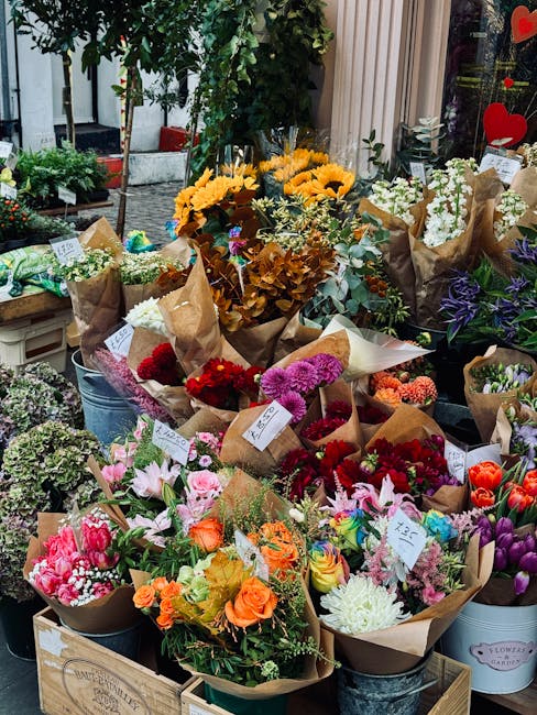 A vibrant display of freshly picked flowers arranged in brown kraft paper wrappings, set outdoors in front of a flower shop. The bouquet arrangements include a variety of colorful blooms such as pink 