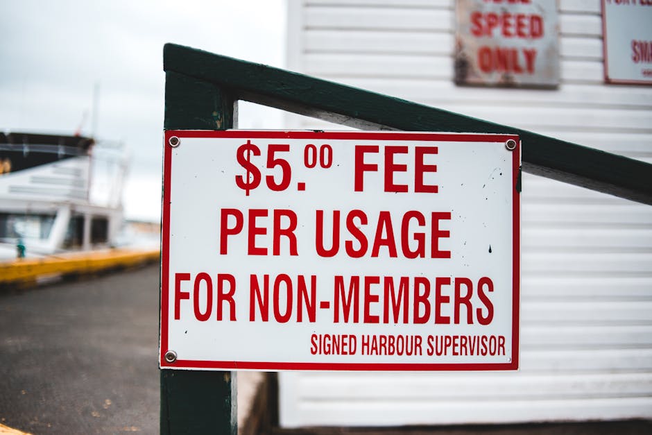 Close-up of a white rectangular sign with bold red lettering stating '$5.00 fee per usage for non-members,' attached to a dark green metal railing at a harbour, with boats docked in the background and