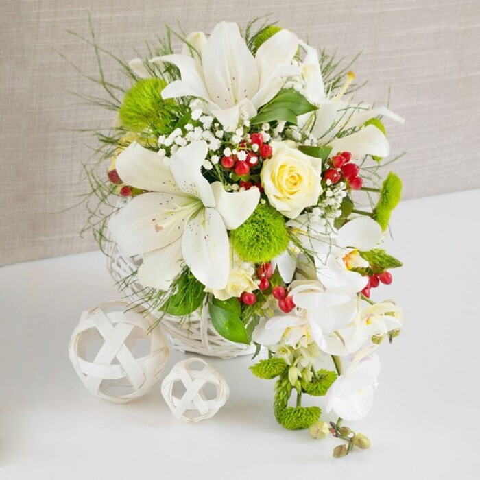 An opulent white and green floral centerpiece, with a graceful cascading element, is displayed on a spotless white surface, as though set for a wedding breakfast in a bright Clapham venue. The arrangement is cradled in a round white woven wicker basket, whose subtle texture enhances the natural, relaxed luxury of the design. Several large Oriental lilies stand proudly at the top and centre, their wide, trumpet-shaped white petals gently curling back to reveal fine dark speckles and prominent stamens. Just above the middle, a single creamy yellow rose sits in perfect bloom, its spiralled petals forming a soft, romantic heart that naturally draws the eye. From the right-hand side, a stream of white Phalaenopsis orchids cascades elegantly downwards, their smooth, flat petals and contrasting yellow throats creating a waterfall effect that gives the whole bouquet a sense of movement-ideal for a head table arrangement in a Clapham Common or Clapham Junction wedding setting. Around these focal flowers, layers of foliage build depth and structure: feathery, wispy greenery; longer arching leaves; and rounded, vivid green pom-pom blooms resembling dianthus or Santini chrysanthemums that lend a velvety, moss-like texture. Fine clusters of tiny white baby's breath spill gently between the stems, softening edges and adding an ethereal haze. Bright red berries, glossy and plump, are nestled throughout, introducing sharp accents of colour that feel both festive and modern. To the left, two decorative white orbs made of interlaced strips rest on the table, their hollow, geometric patterns echoing the basket's weave. The neutral, lightly textured beige background keeps all attention on the flowers, creating an impression of calm, refined celebration, like a bespoke design prepared by a local Clapham florist for a sophisticated city wedding or celebratory dinner.